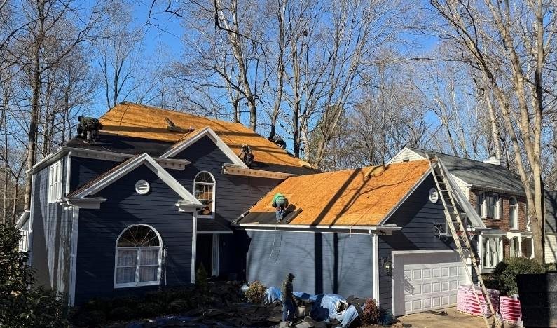 During: the house stripped to bare orange roof decking with the crew working on it and materials stacked in the drive.