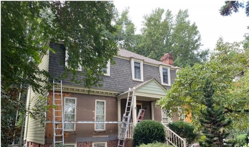 Before: a two-story home in dark brown shingle siding with ladders leaning against the gable.