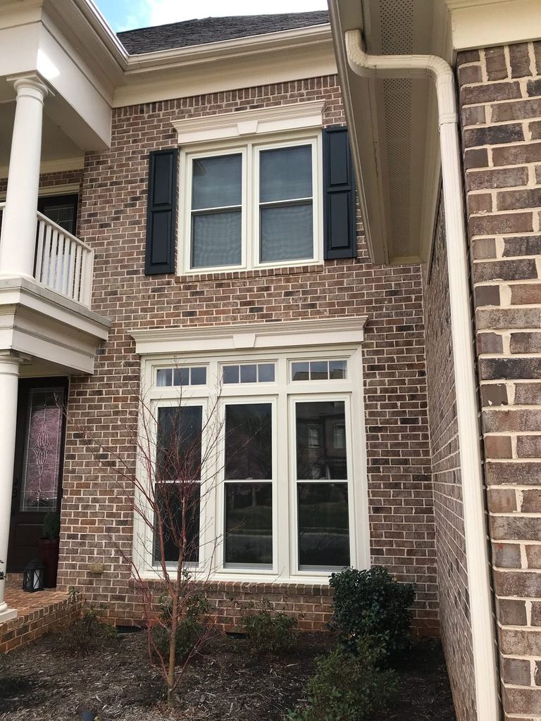 New white Simonton replacement windows in a Ballantyne brick two-story, black shutters above and a white portico column to the left.